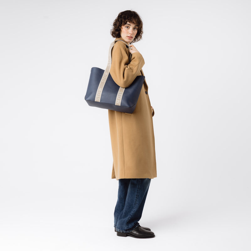 Model wearing STOW London's Nastro Tote bag in Navy pebbled leather with beige nastro handles featuring 'STOW' branding on her shoulder on a white background