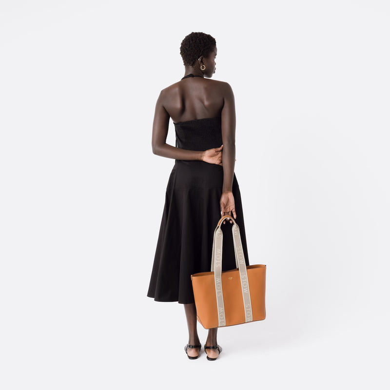Model holding STOW London's Nastro Tote bag in Earth Tan pebbled leather with beige nastro handles featuring 'STOW' branding on a white background