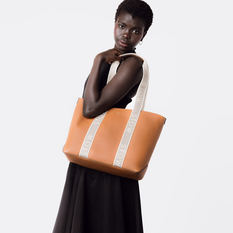 Model holding STOW London's Nastro Tote bag in Earth Tan pebbled leather on her shoulder with beige nastro handles featuring 'STOW' branding on a white background
