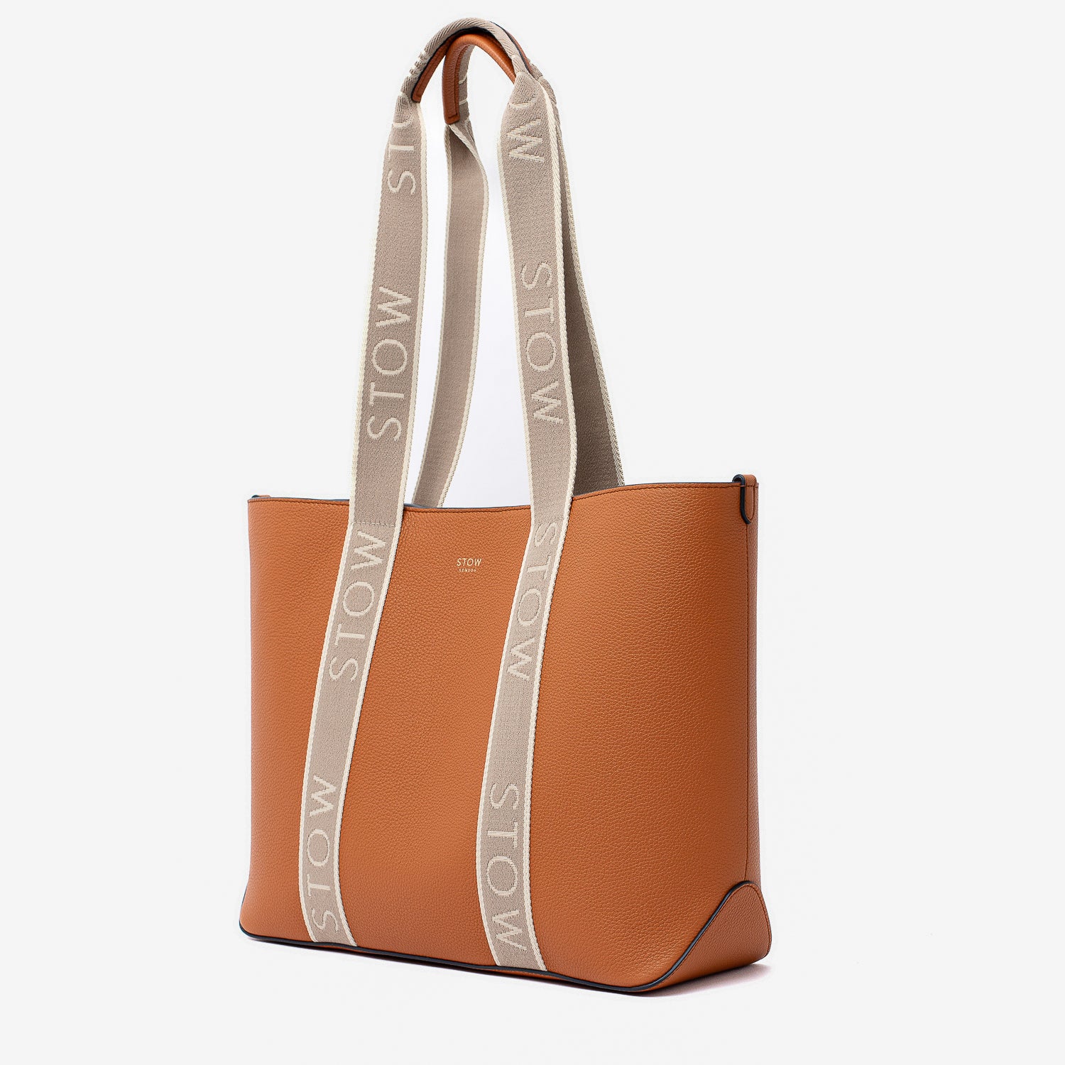 Off-axis view of STOW London's Nastro Tote bag in Earth Tan pebbled leather with beige nastro handles featuring 'STOW' branding on a light grey background