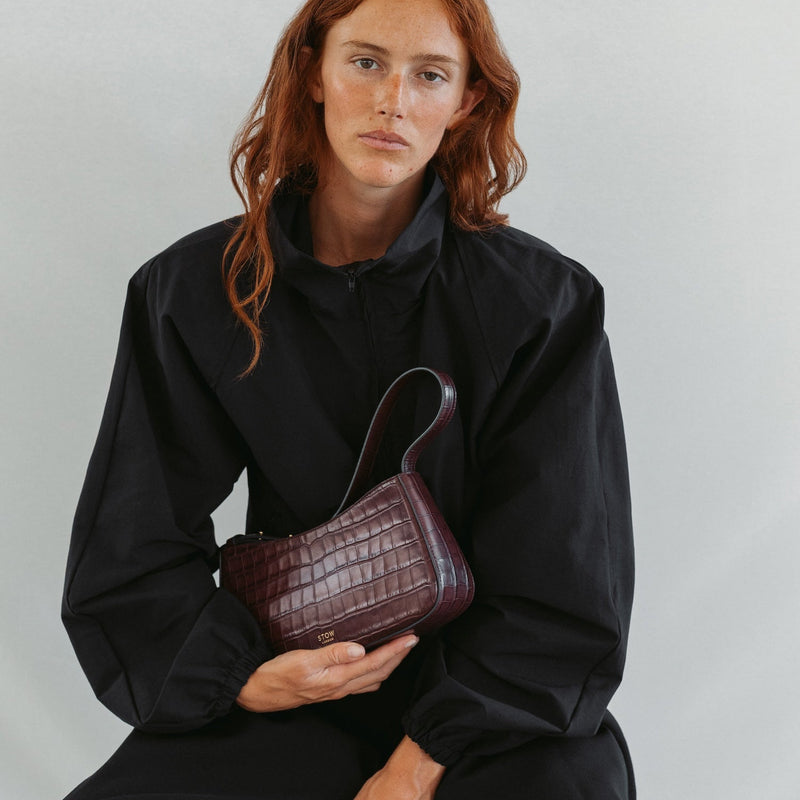Model holding STOW London's Post Shoulder bag in Burgundy Embossed Croc leather featuring gold-coated stainless steel hardware 