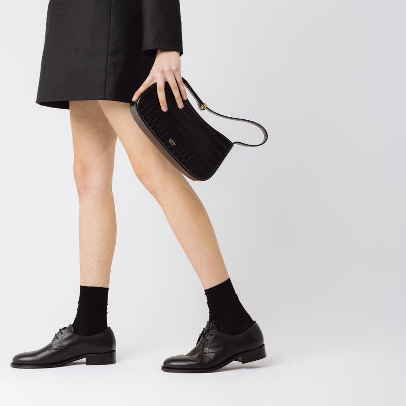 Model holding STOW London's Post Shoulder bag in Black Embossed Croc leather in her hand at her side featuring gold-coated stainless steel hardware on a white background