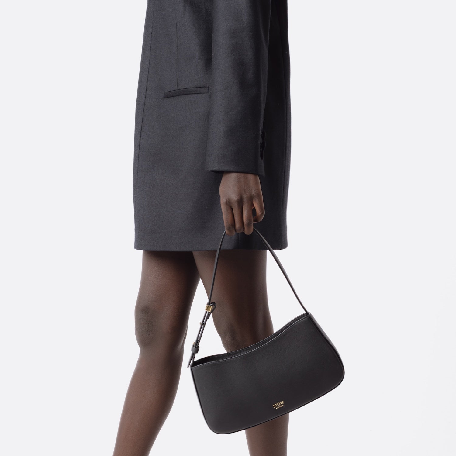 Model holding STOW London's Post Shoulder bag in Black smooth leather at her side featuring gold-coated stainless steel hardware on a white background