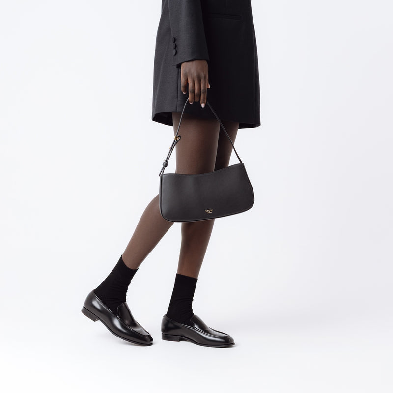 Model holding STOW London's Post Shoulder bag in Black smooth leather at her side featuring gold-coated stainless steel hardware on a white background