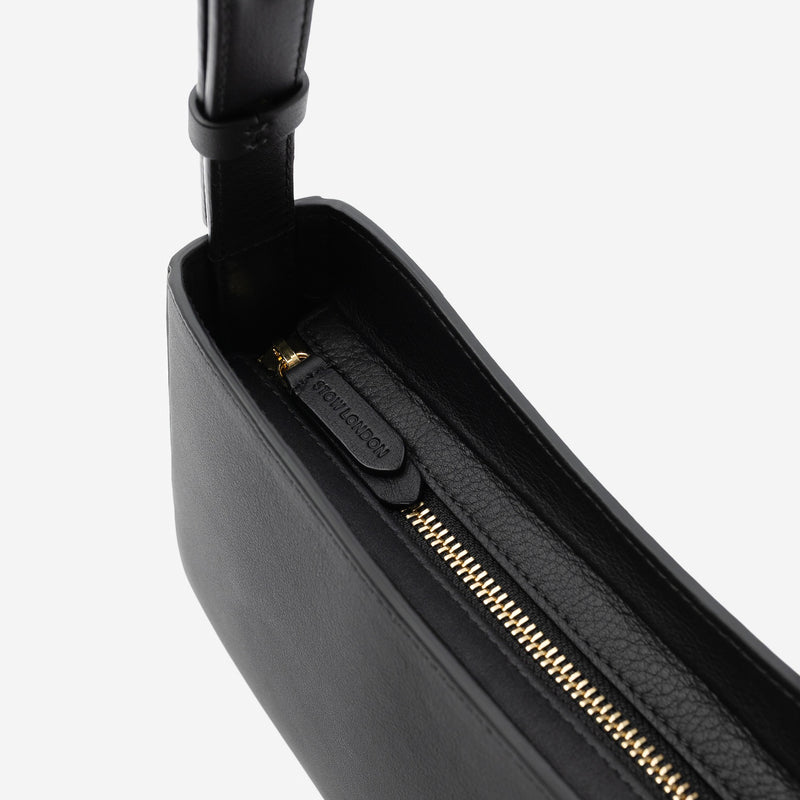 Close up of the gold zip closure and STOW London embossed pull tabs on Stow London's Post Shoulder in Black smooth leather on a light grey background 
