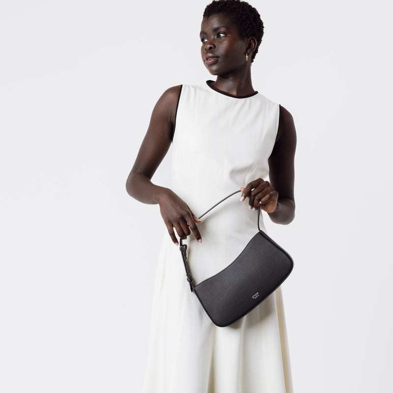 Model holding STOW London's Post Shoulder bag in Black smooth leather at her front featuring silver hardware on a white background