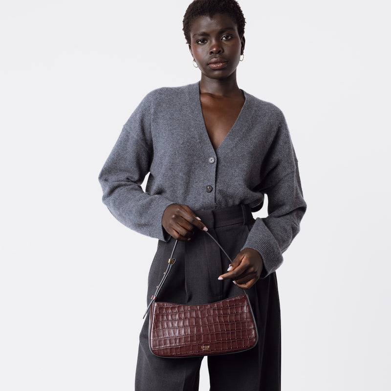 Model holding STOW London's Post Shoulder bag in Burgundy Embossed Croc leather at her front featuring gold-coated stainless steel hardware on a white background