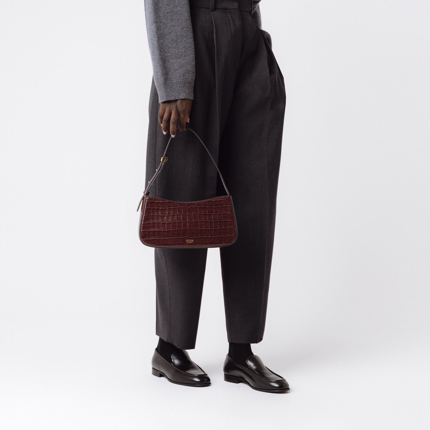 Model holding STOW London's Post Shoulder bag in Burgundy Embossed Croc leather in front of her featuring gold-coated stainless steel hardware on a white background