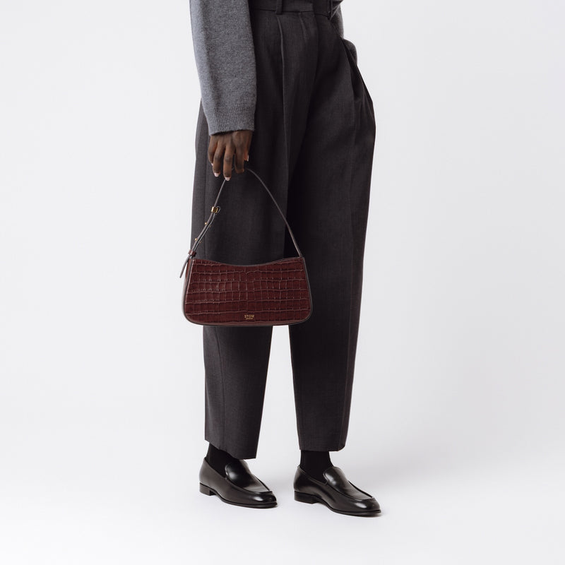 Model holding STOW London's Post Shoulder bag in Burgundy Embossed Croc leather in front of her featuring gold-coated stainless steel hardware on a white background