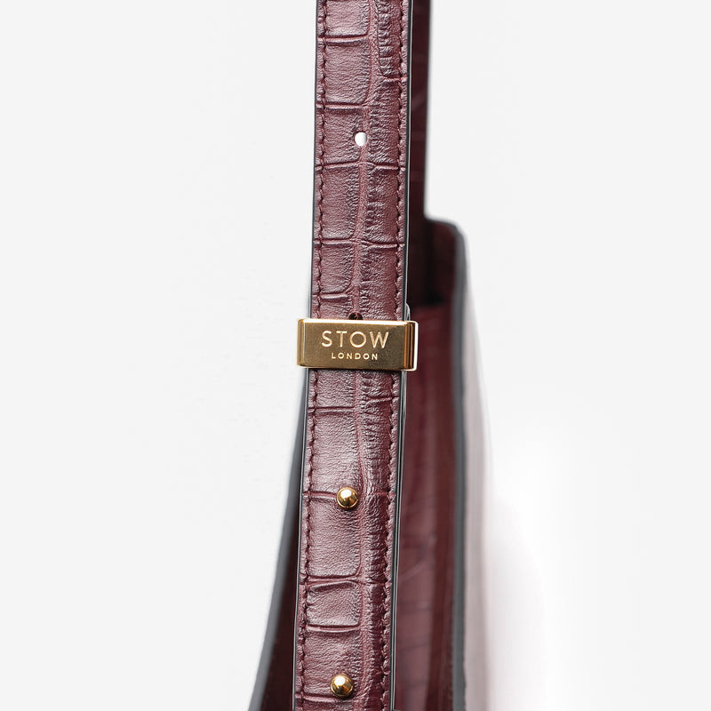 Close up of the gold hardware on the adjustable strap on Stow London's Post Shoulder in Burgundy Embossed Croc leather on a light grey background 