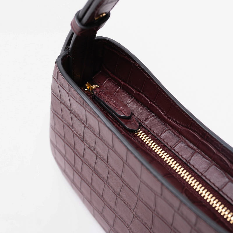 Close up of the gold zip closure and STOW embossed pull tabs on Stow London's Post Shoulder in Burgundy Embossed Croc leather on a light grey background featuring gold-coated stainless steel hardware
