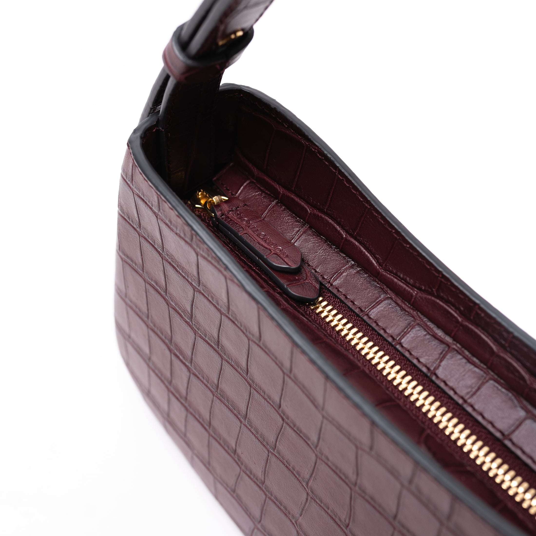 Close up of zipper and pull tab on STOW London's Post Shoulder bag in Burgundy Embossed Croc leather featuring gold-coated stainless steel hardware on a white background