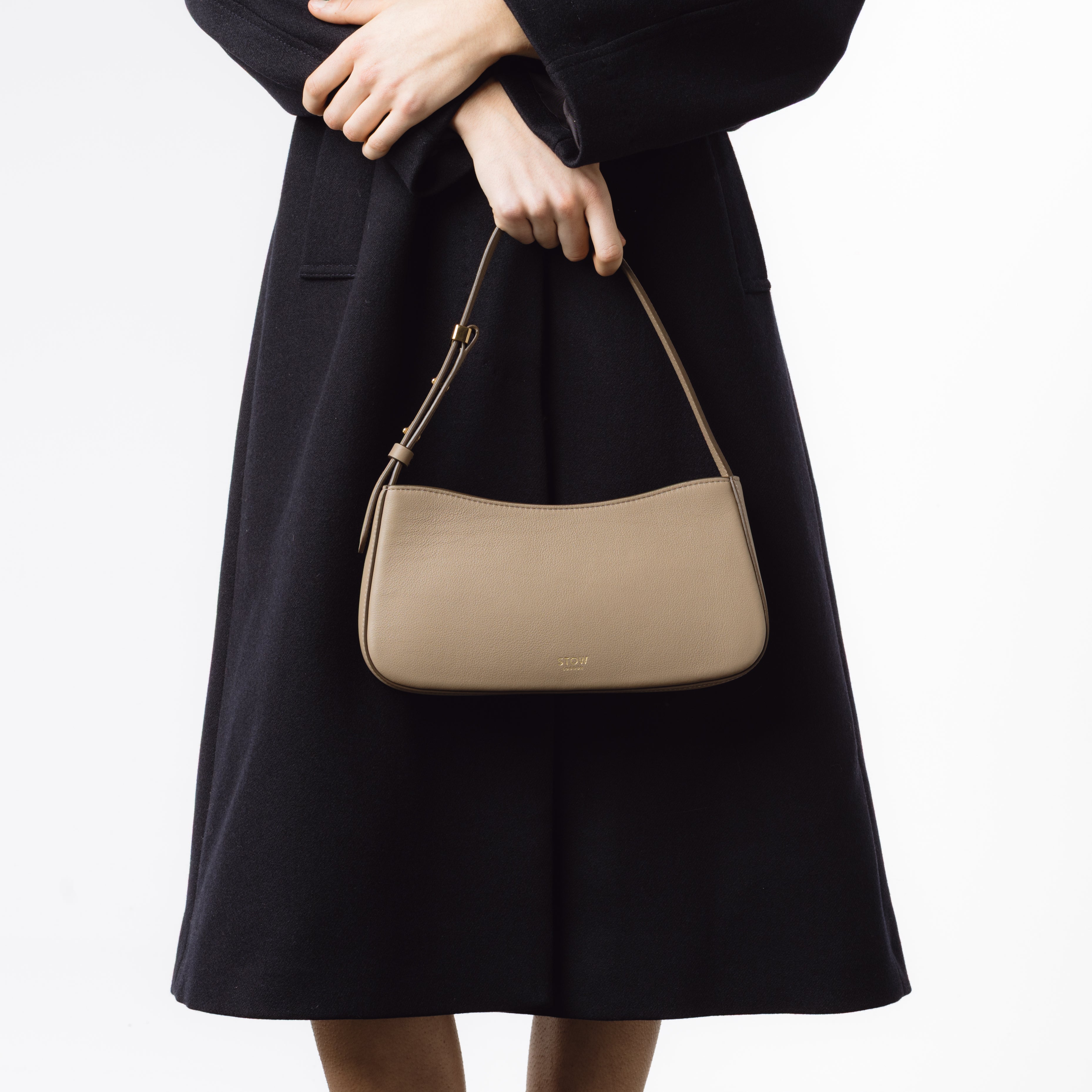 Model holding STOW London's Post Shoulder bag in Warm Taupe smooth leather at her front featuring gold-coated stainless steel hardware on a white background
