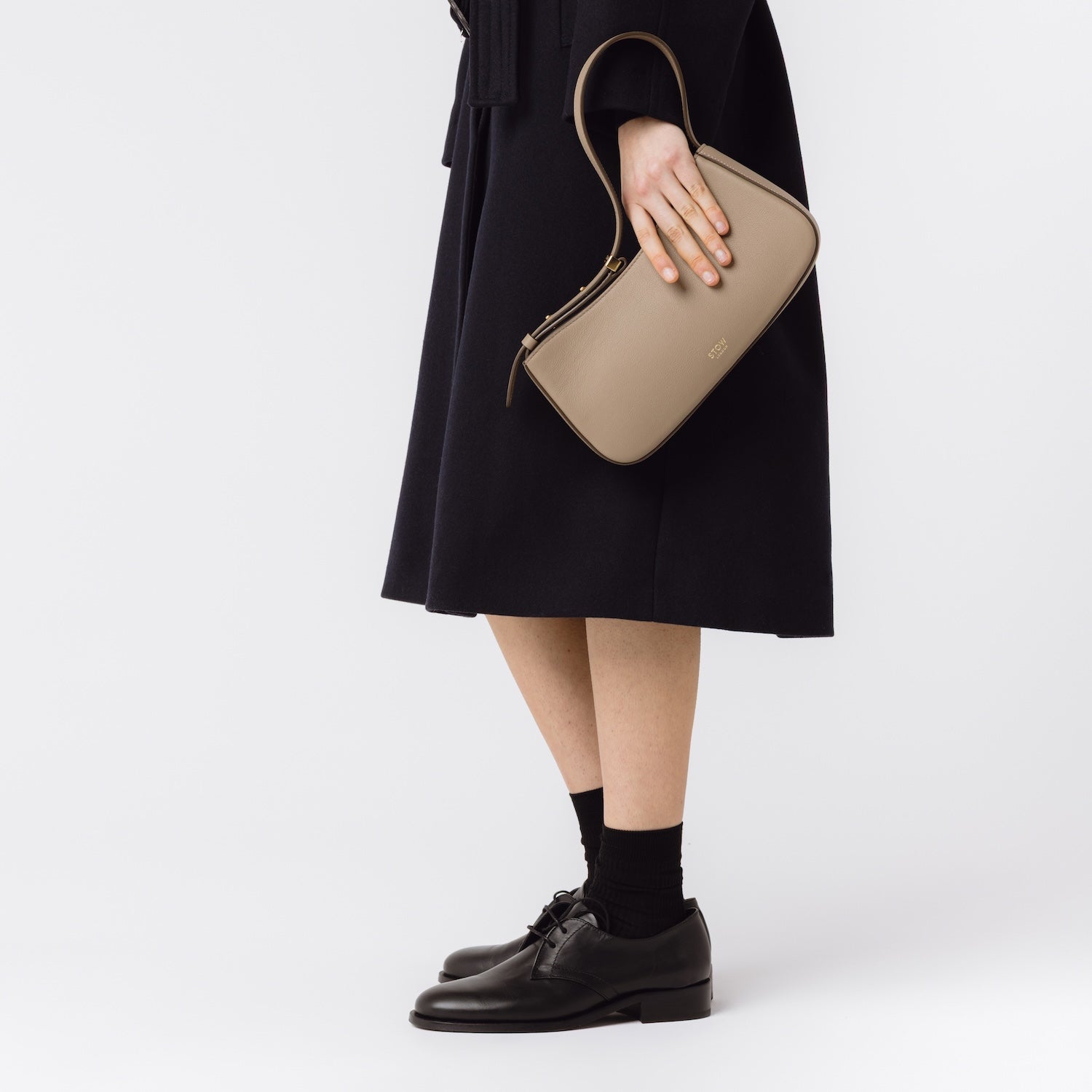 Model holding STOW London's Post Shoulder bag in Warm Taupe smooth leather in her hand at her side featuring gold-coated stainless steel hardware on a white background