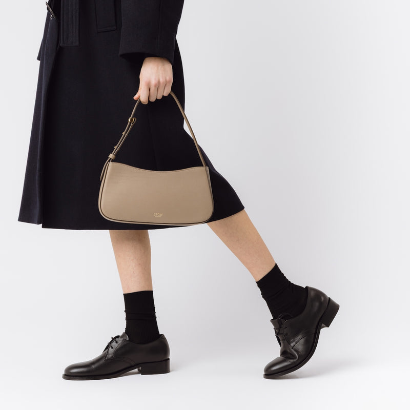 Model carrying STOW London's Post Shoulder bag in Warm Taupe smooth leather at her side featuring gold-coated stainless steel hardware on a white background