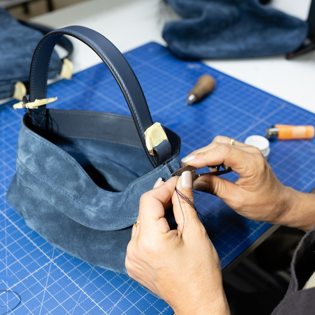 A close-up of a person's hands crafting a dark blue suede handbag on a blue cutting mat, with leatherworking tools visible in the background.