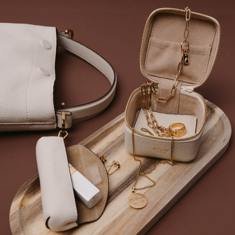STOW London's Hester Jewellery Case in Spring Moon pebbled leather filled with gold jewellery, alongside the Spring Moon Lipstick case and Midi Curve on a wooden platter. 