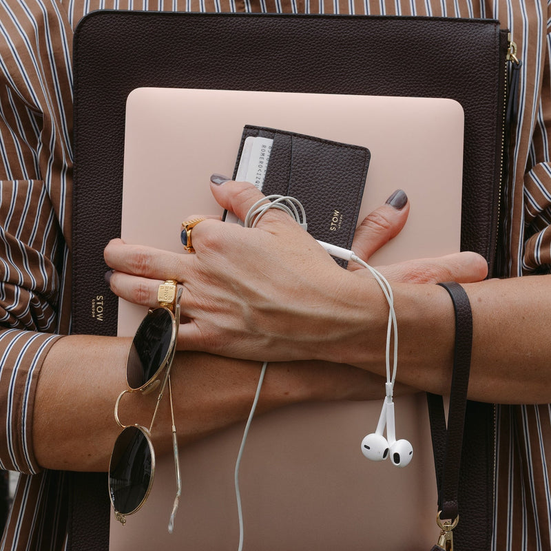 Woman holding STOW London's Leather Sleeve and Cardholder in Espresso pebbled leather, alongside a peach laptop, sunglasses and earphones. 