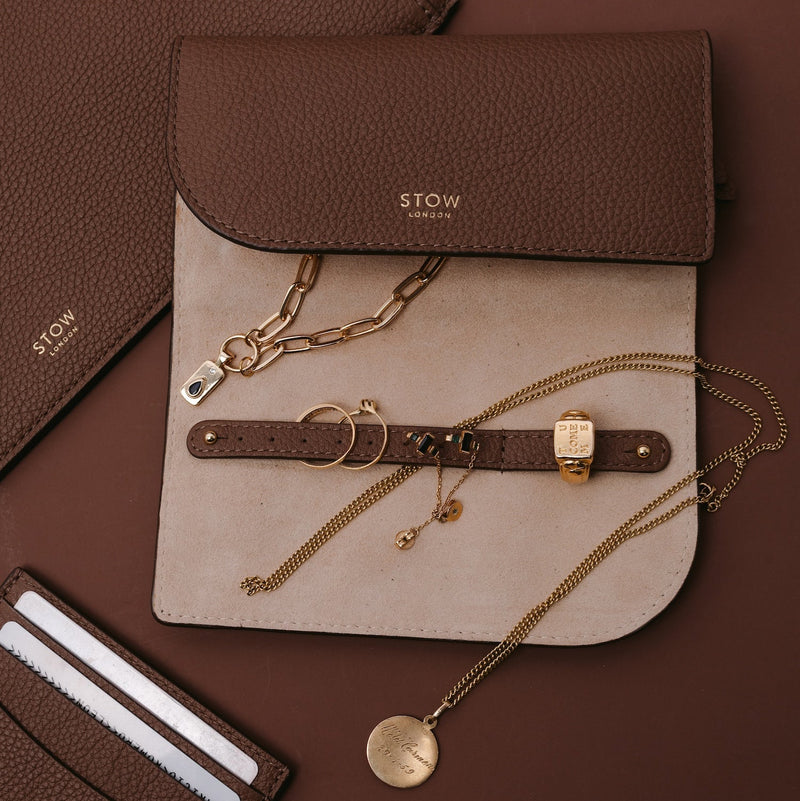 STOW London's Jewellery Roll in Mocha pebbled leather filled with gold necklaces, rings and earrings alongside the Mocha cardholder and leather sleeve on a brown background 