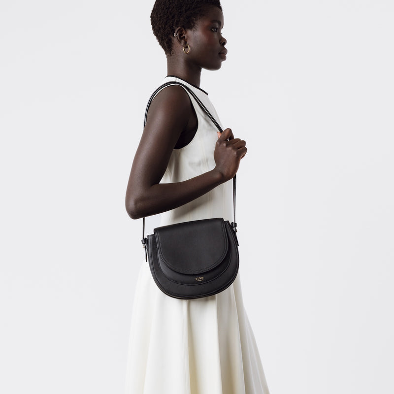 Model wearing STOW London's Saddle bag in Black smooth leather on her shoulder by the detachable and adjustable  crossbody strap featuring gold-coated stainless steel hardware on a white background