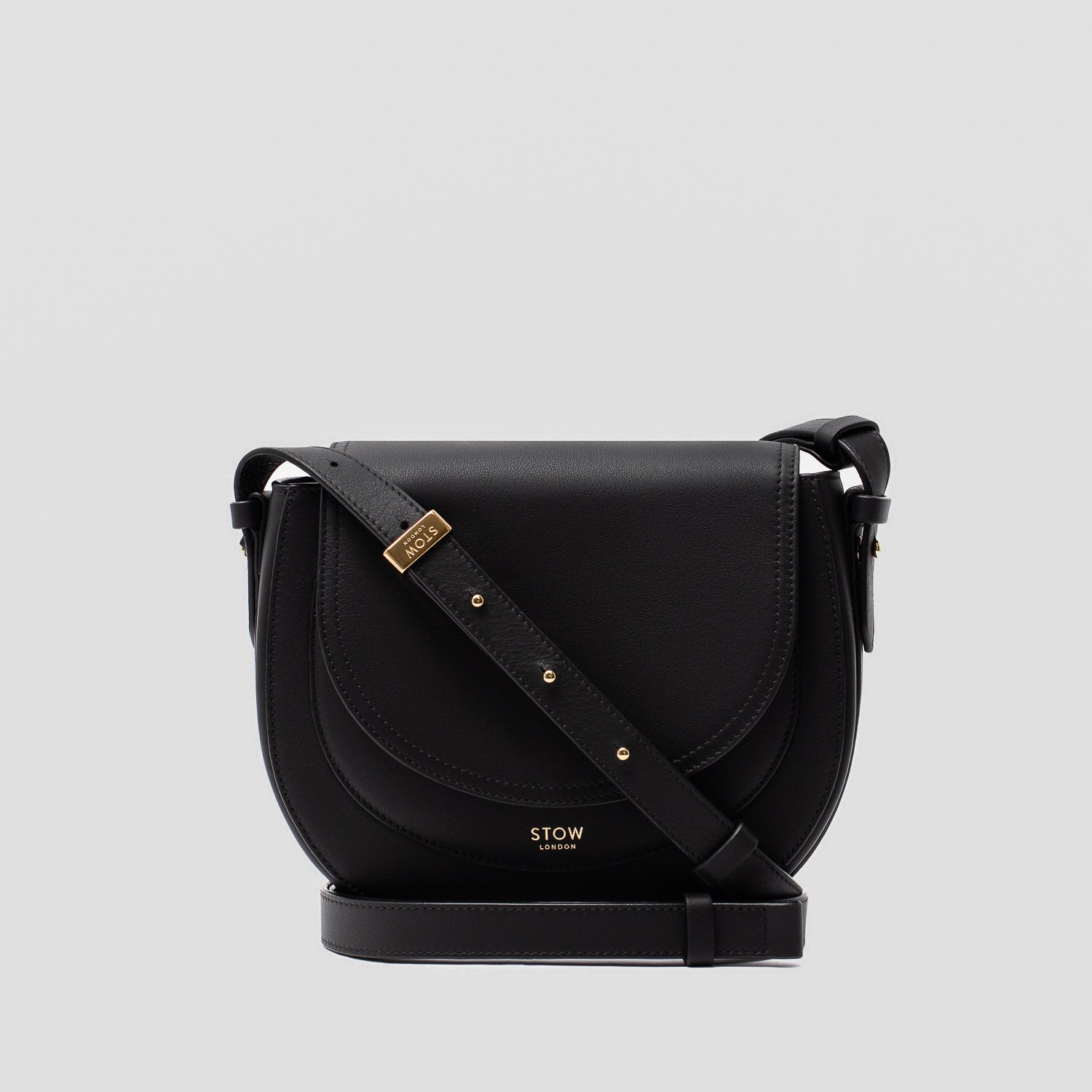 STOW London's Saddle bag in Black smooth leather and detachable crossbody strap with gold-coated stainless steel hardware on a light grey background