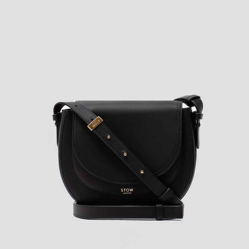 STOW London's Saddle bag in Black smooth leather and detachable crossbody strap with gold-coated stainless steel hardware on a light grey background