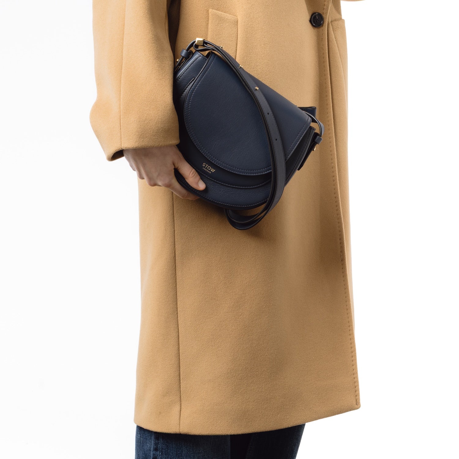 Model holding STOW London's Saddle bag in Navy smooth leather in her hand at her side featuring gold-coated stainless steel hardware and the crossbody detachable strap on a white background