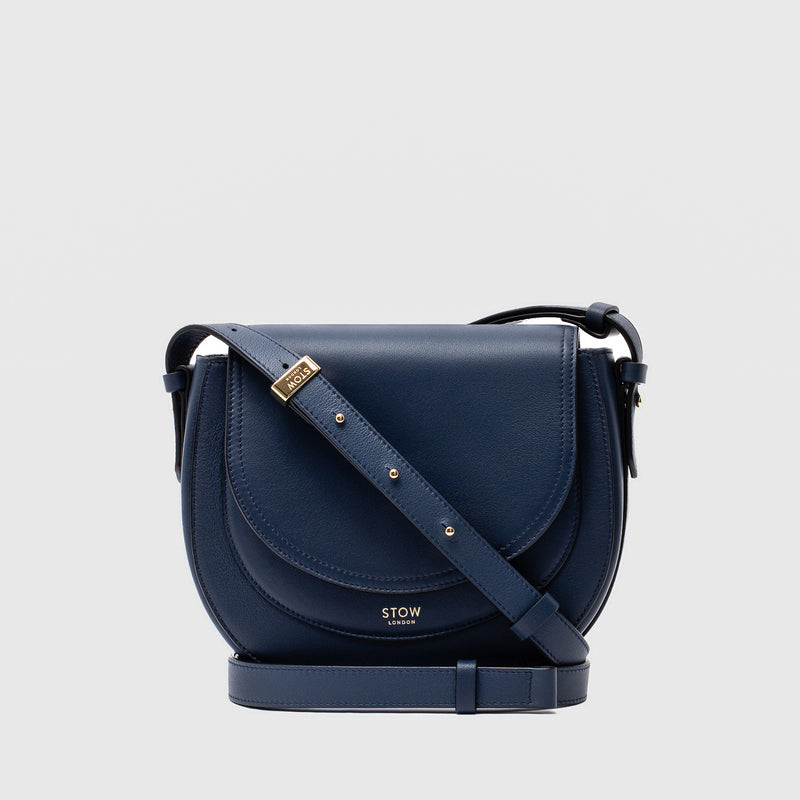 STOW London's Saddle bag in Navy smooth leather and detachable crossbody strap featuring gold-coated stainless steel hardware on a light grey background