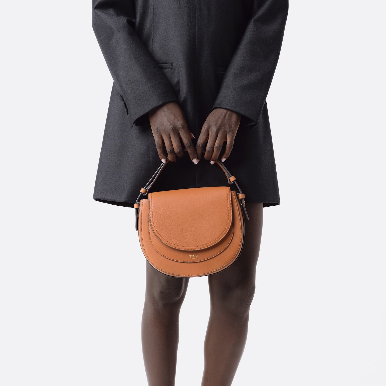 STOW London's Saddle bag in Earth Tan smooth leather held by top handle at model's front on a white background