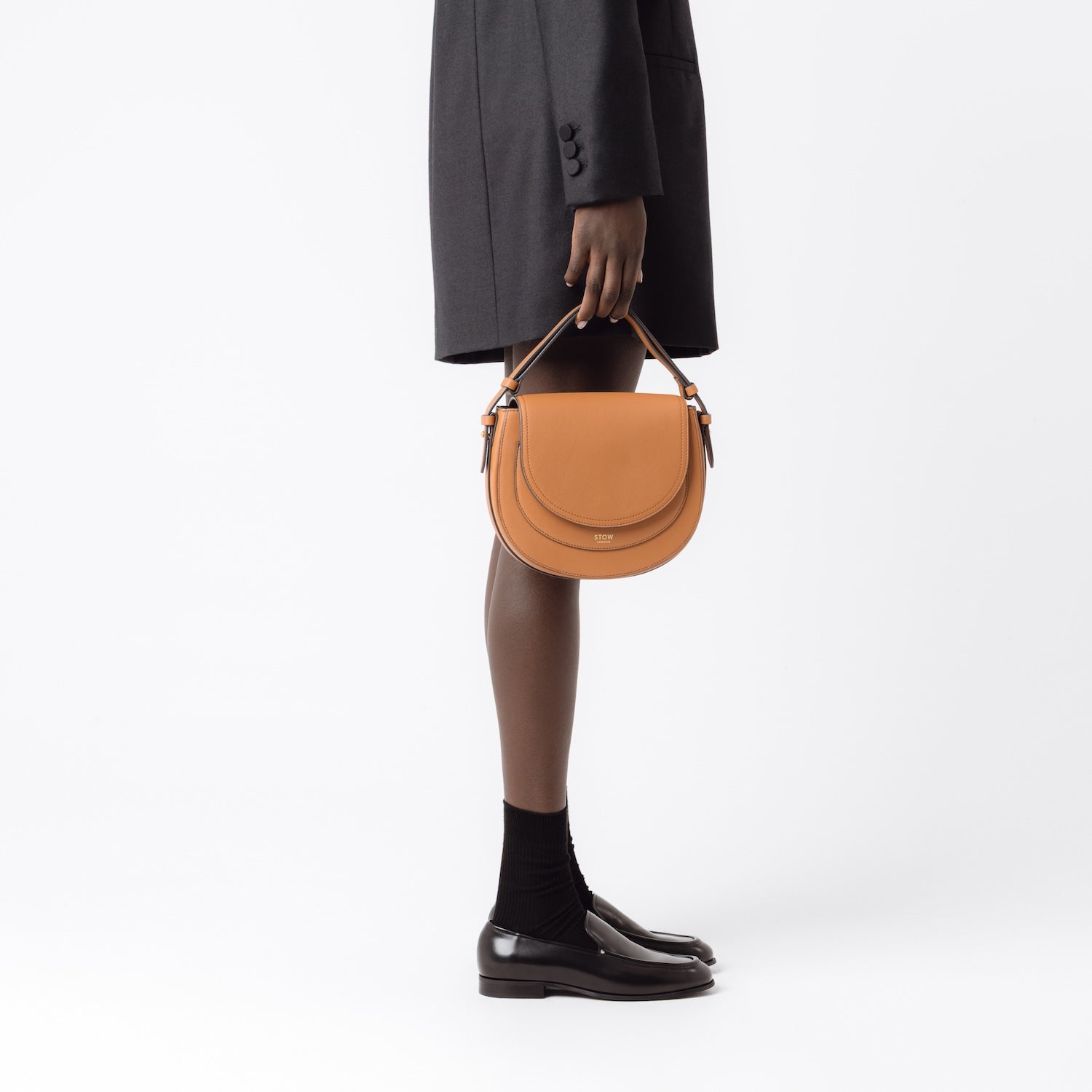 Model holding STOW London's Saddle bag in Earth Tan smooth leather by its top handle at her side featuring gold-coated stainless steel hardware on a white background