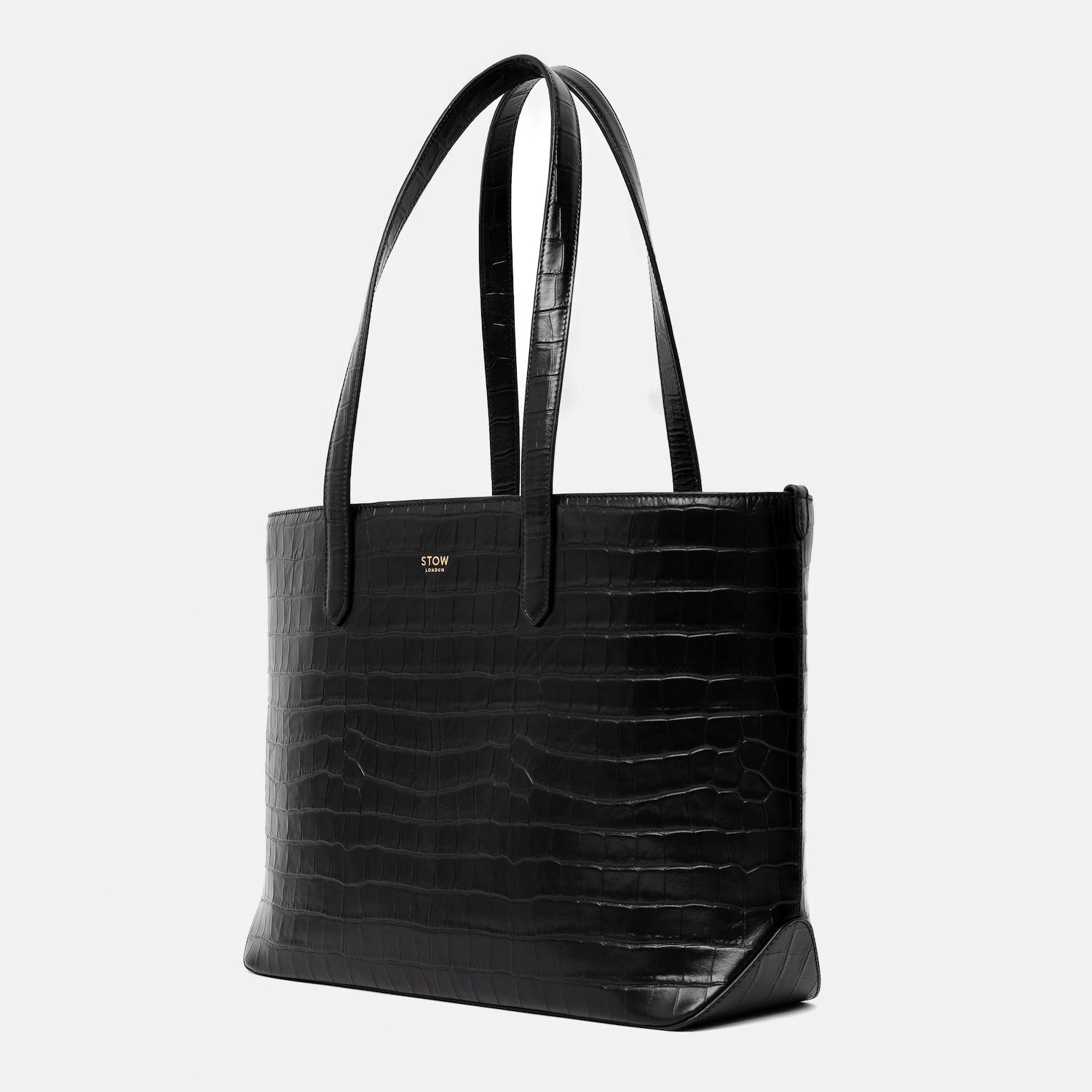 Off-axis view of Stow London's Everyday Tote Bag in Black Embossed Croc leather on a light grey background featuring gold STOW logo