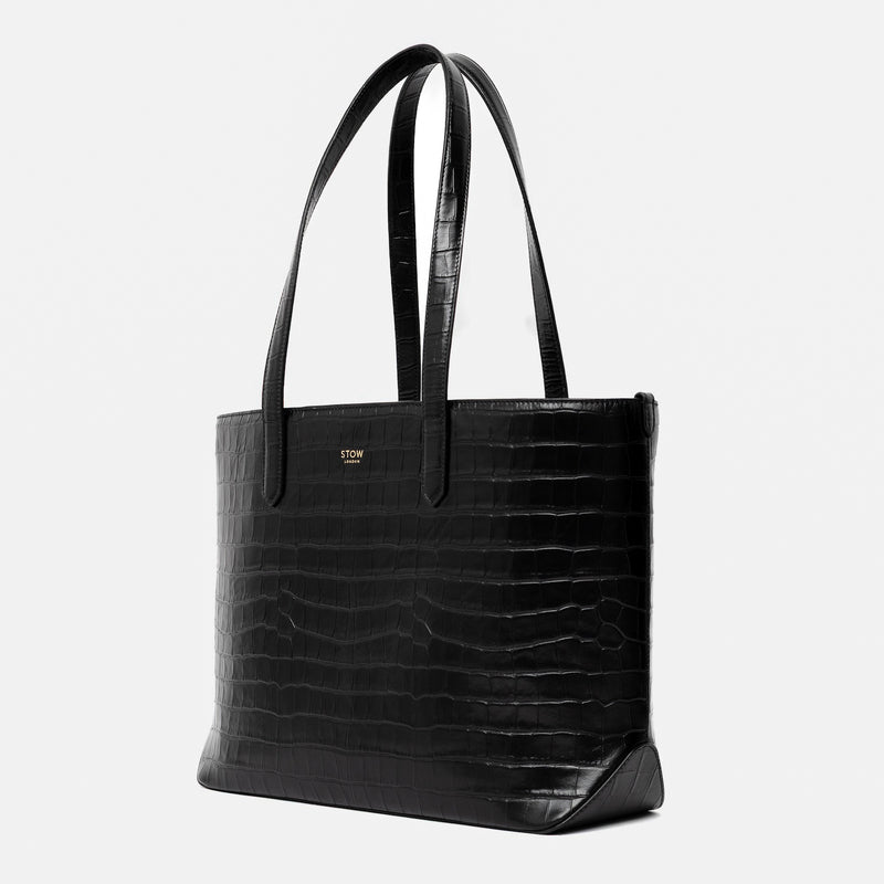 Off-axis view of Stow London's Everyday Tote Bag in Black Embossed Croc leather on a light grey background featuring gold STOW logo