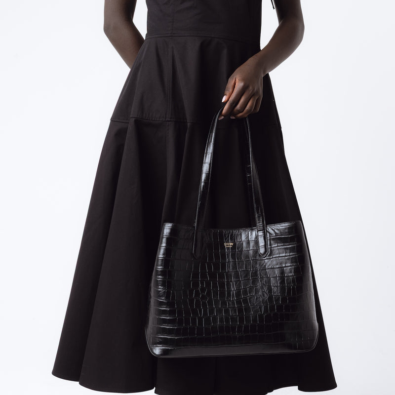 Model holding Stow London's Everyday Tote Bag in Black Embossed Croc leather on a white background featuring gold STOW logo