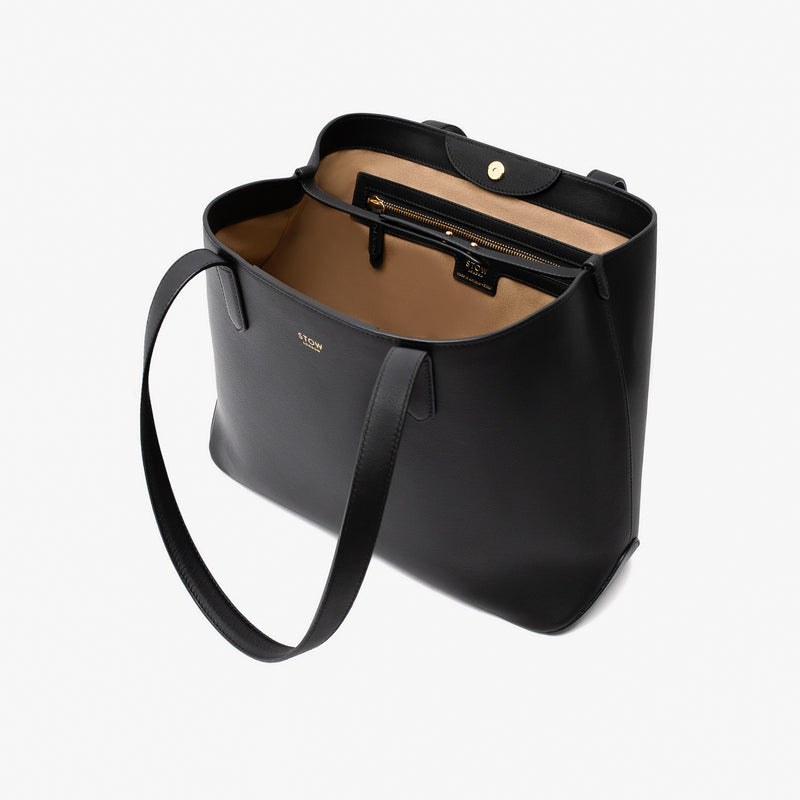 Stow London's Everyday Tote Bag in Black smooth leather on a light grey background with magnets open showing light beige cotton interior featuring the leather strap, zip pocket and magnetic closure