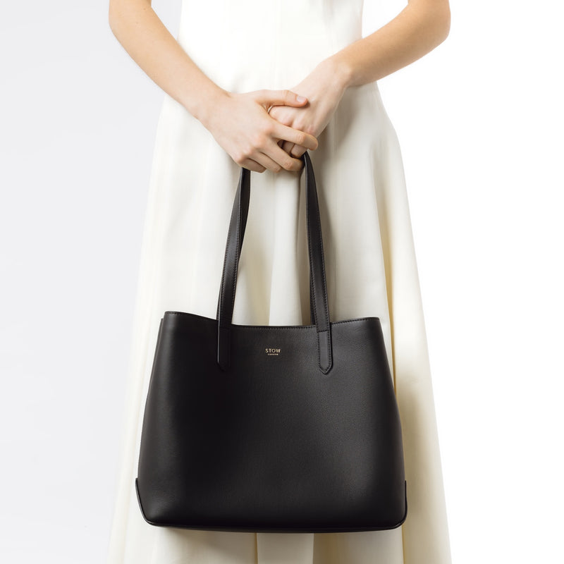 Model holding Stow London's Everyday Tote Bag in Black smooth leather on a light grey background featuring gold STOW logo
