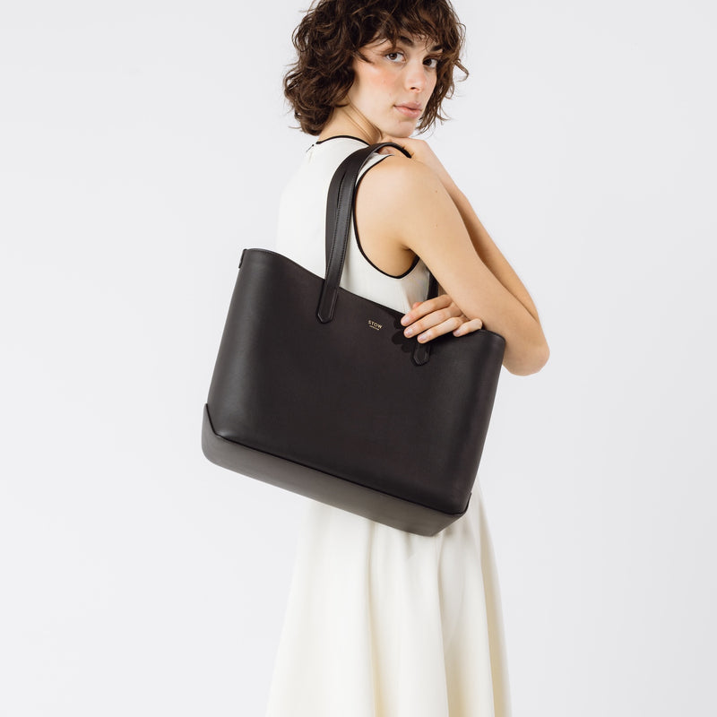 Model wearing Stow London's Everyday Tote Bag in Black smooth leather on her shoulder on a light grey background featuring gold STOW logo