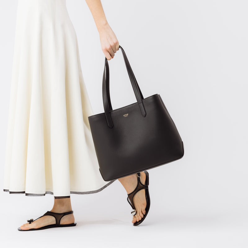 Model holding Stow London's Everyday Tote Bag in Black smooth leather at he side on a whitebackground featuring gold STOW logo