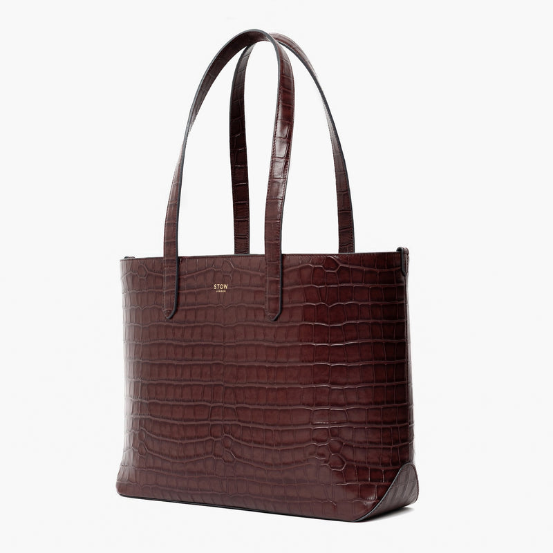 Off-axis view of Stow London's Everyday Tote Bag in Burgundy Embossed Croc leather on a light grey background featuring gold STOW logo