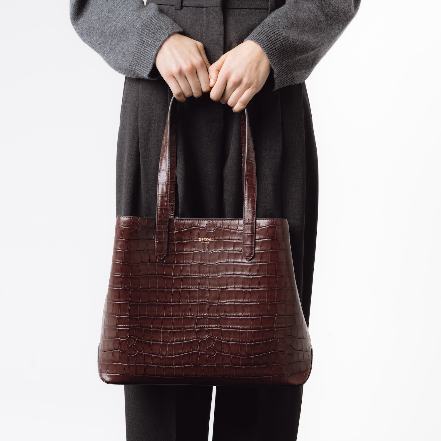 Model holding Stow London's Everyday Tote Bag in Burgundy Embossed Croc leather by the handles at her front featuring gold hardware on a white background