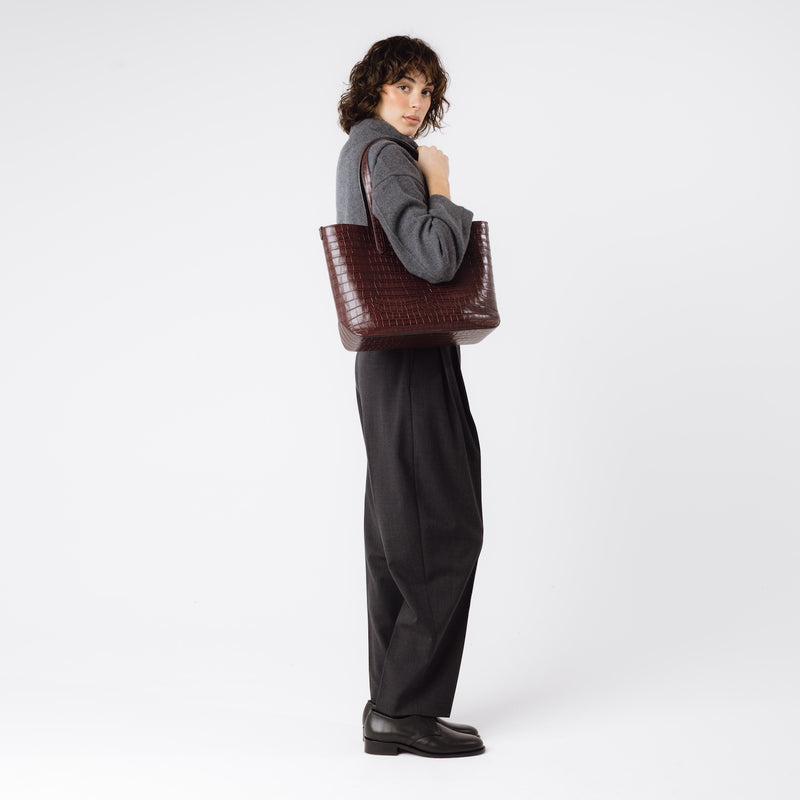 Model wearing Stow London's Everyday Tote Bag in Burgundy Embossed Croc leather on her shoulder featuring gold hardware on a white background