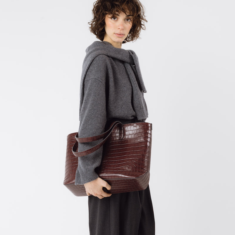 Model holding Stow London's Everyday Tote Bag in Burgundy Embossed Croc leather at her side featuring gold hardware on a white background