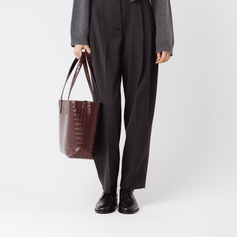 Model holding Stow London's Everyday Tote Bag in Burgundy Embossed Croc leather at her side on a white background