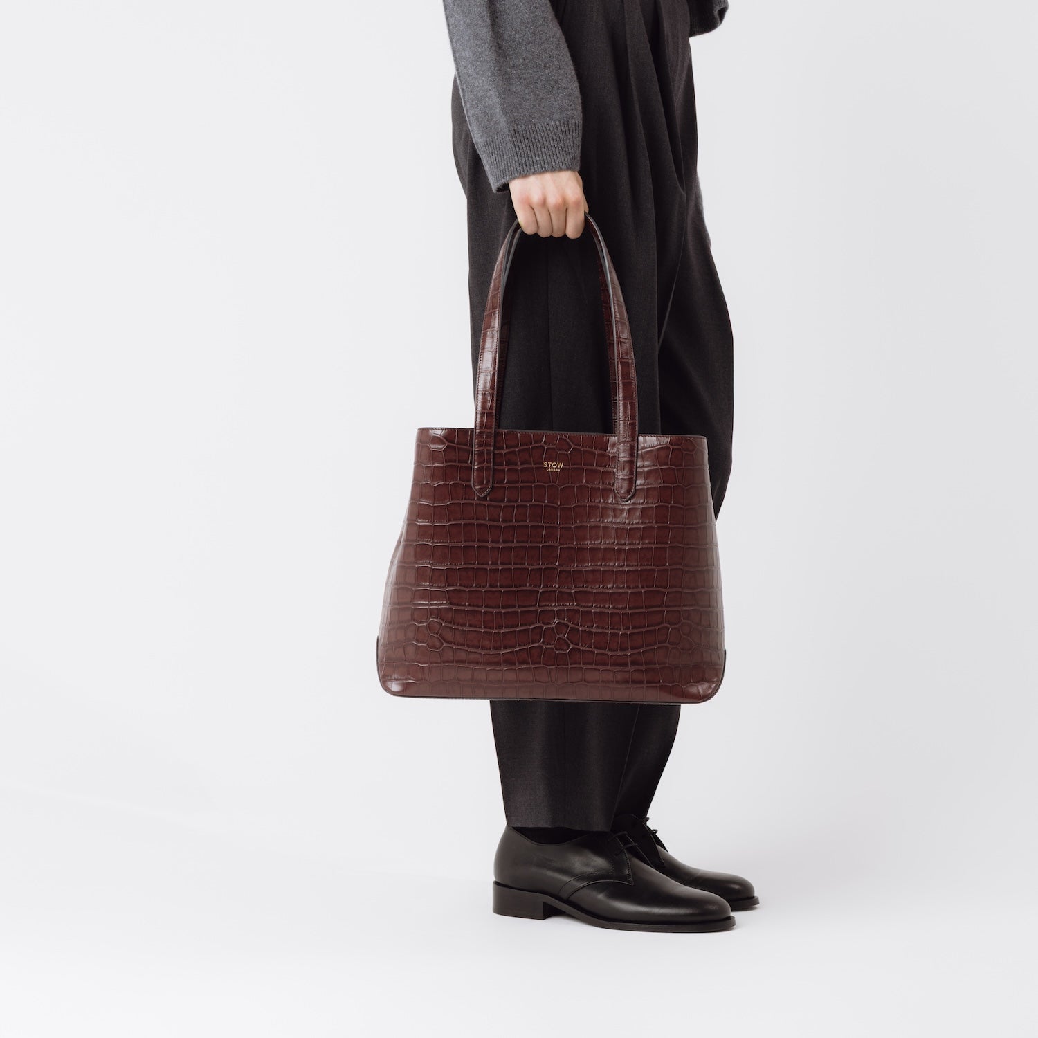 Model holding Stow London's Everyday Tote Bag in Burgundy Embossed Croc leather by the handles at her side featuring gold hardware on a white background