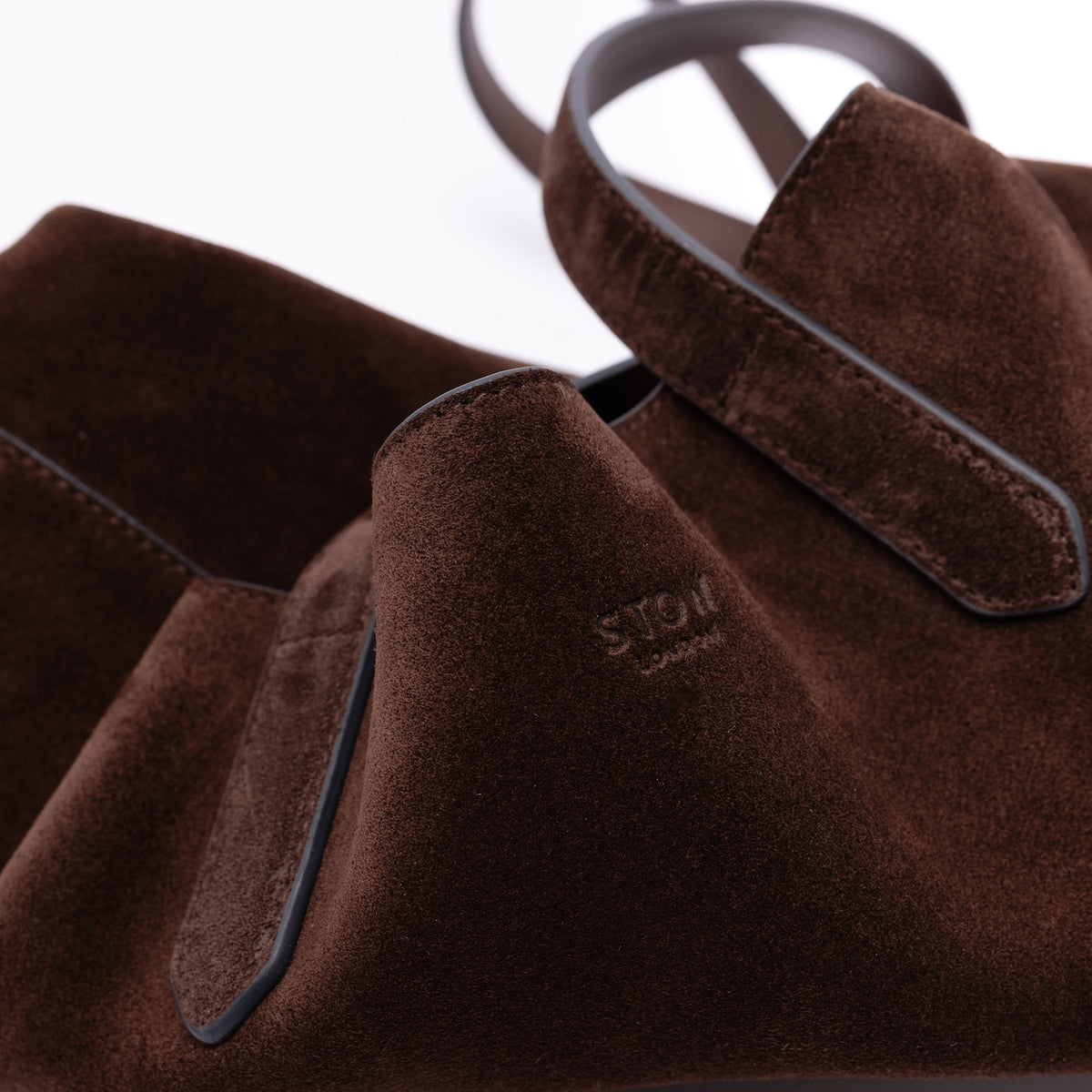 Close up of the small embossed logo on STOW London's Everyday Tote bag in Chocolate suede on a light grey background 