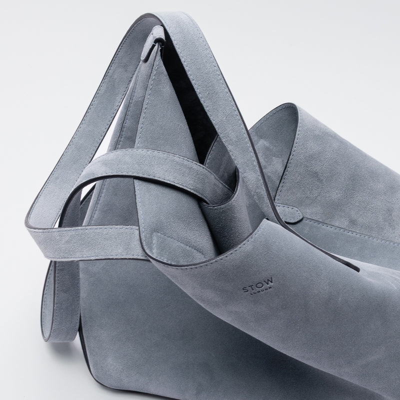 Close up of STOW London's Everyday Tote bag in Misty Blue suede on a light grey background featuring a small STOW London logo embossed on the front panel 