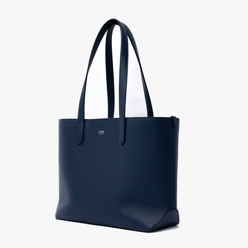 Off-axis view of Stow London's Everyday Tote Bag in Navy smooth leather on a light grey background featuring gold STOW logo