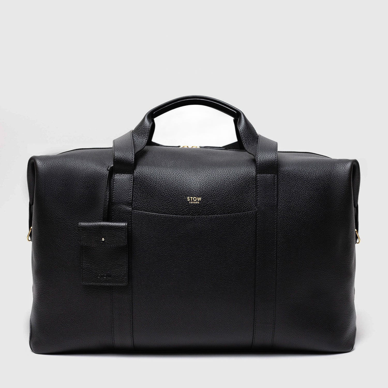 STOW London's Weekend bag in Black pebbled leather featuring detachable pouch and gold STOW London logo on front panel on a light grey background