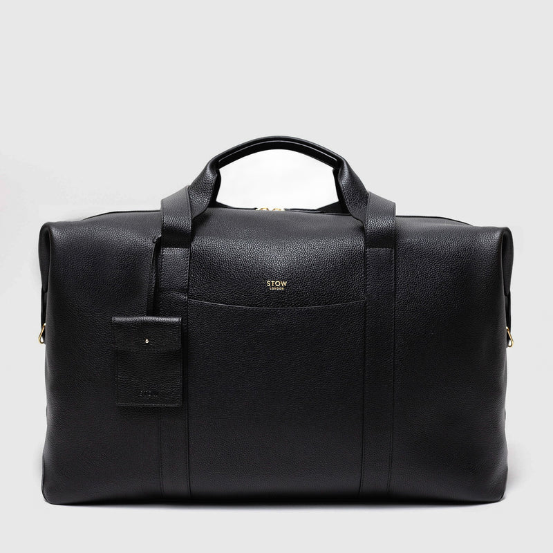 STOW London's Weekend bag in Black pebbled leather featuring detachable pouch and gold STOW London logo on front panel on a light grey background