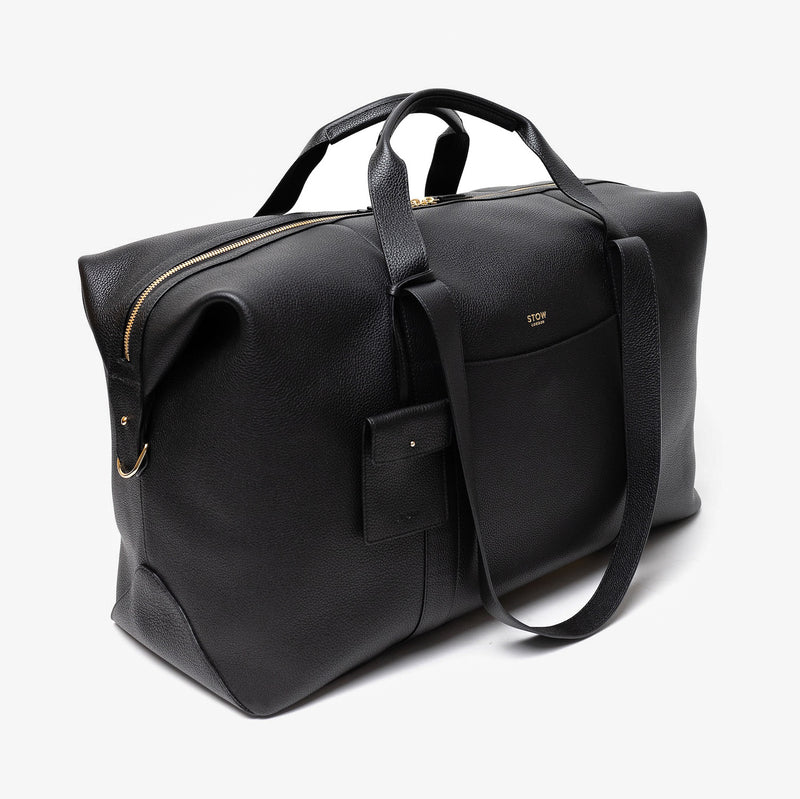Off-axis view of STOW London's Weekend bag in Black pebbled leather featuring detachable pouch and gold-coated stainless steel hardware on a light grey background
