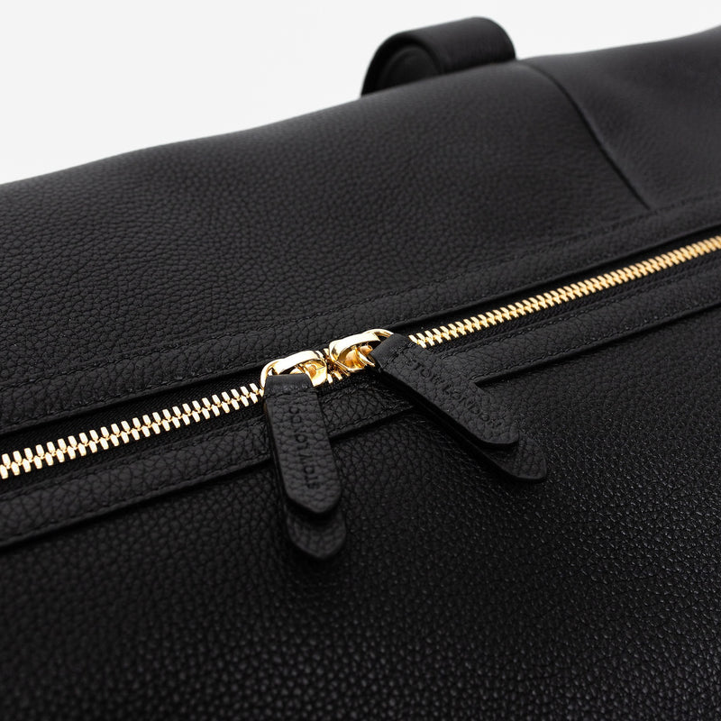 Close up of the gold zip closure and STOW London embossed pull tabs on STOW London's Weekend bag in Black pebbled leather on a light grey background